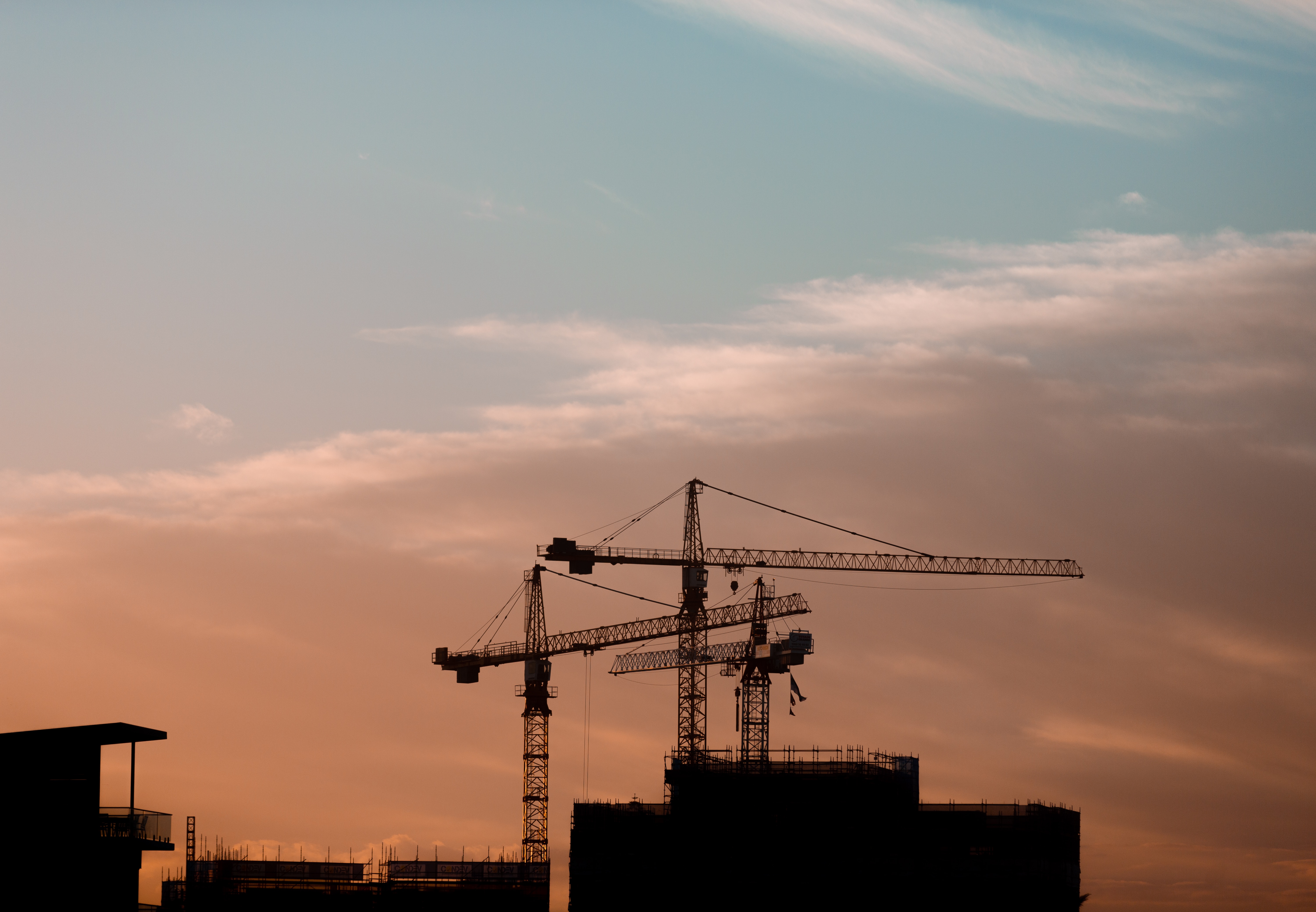 Two cranes on top of buildings in front of a pink sunset