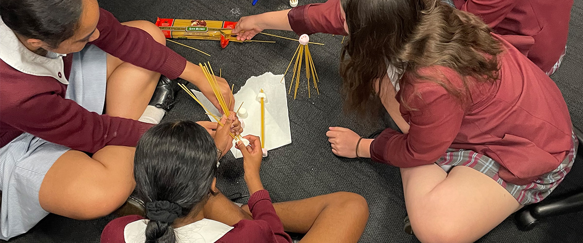 Three girls sitting in a circle making structures with long thin pasta and marshmallows