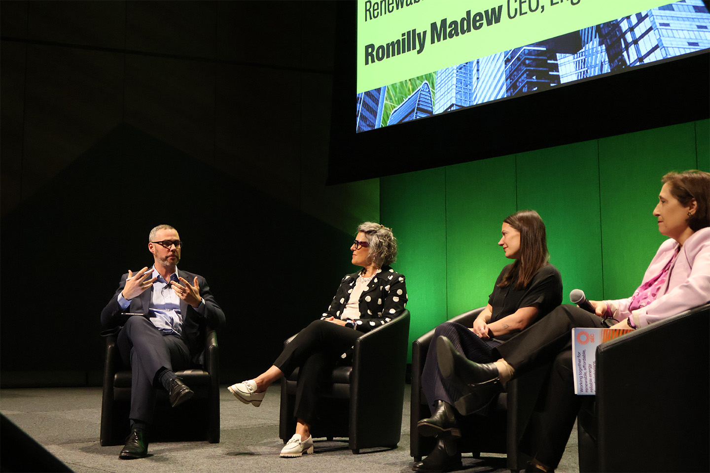 Romilly Madew sitting on a chair, on a stage, alongside two other women and a man
