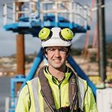 Headshot of Jarrod in front of a construction site wearing a yellow safety vest, yellow earmuffs and a white helmet